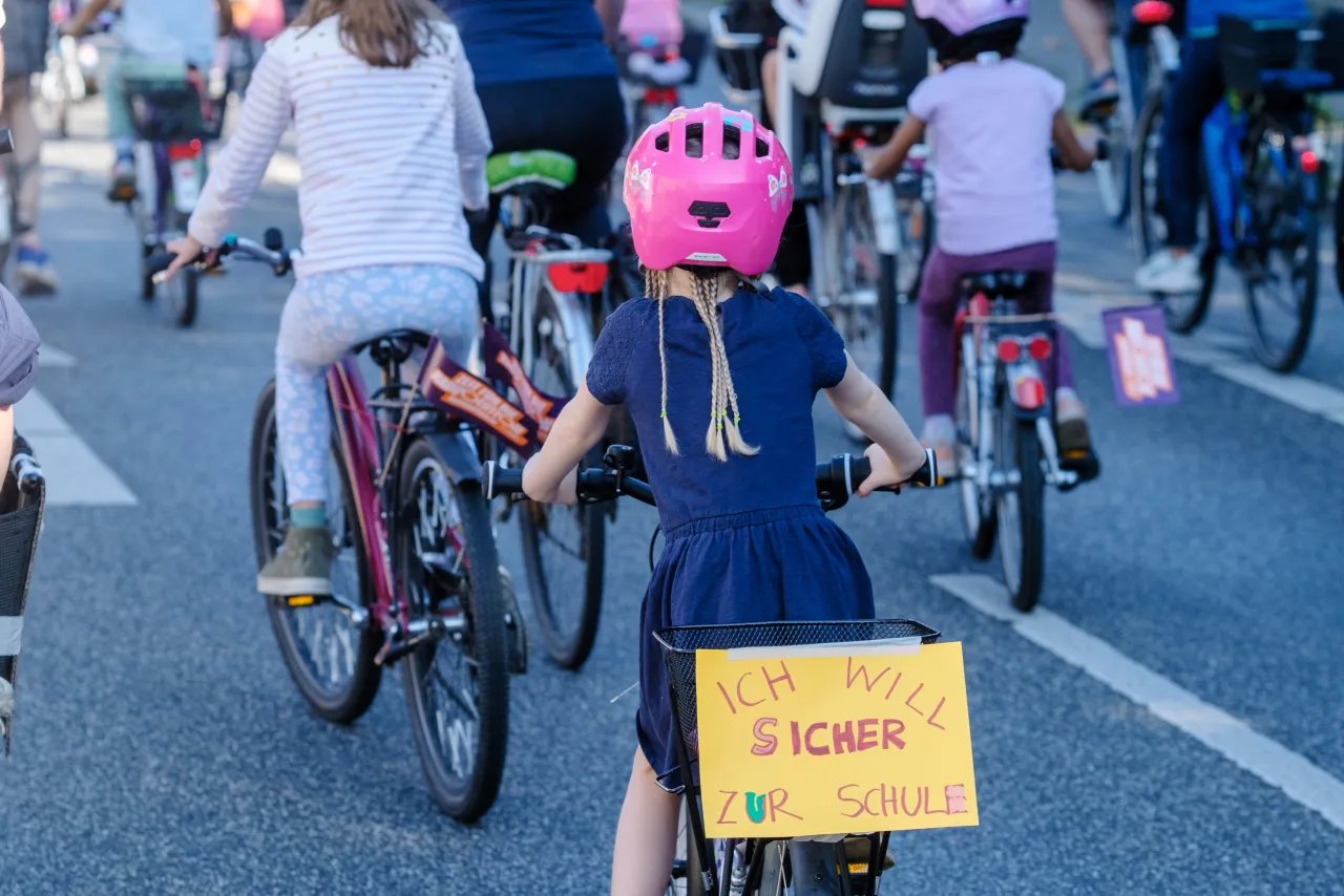 Eine Gruppe von Kindern fährt mit Fahrrädern auf einer Straße, ein Mädchen in einem dunkelblauen Kleid mit pinkem Helm hat ein Schild am Fahrradkorb mit der Aufschrift "Ich will sicher zur Schule".